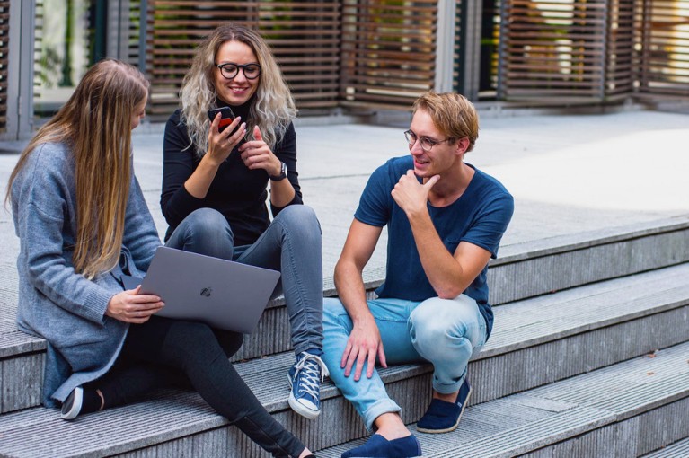 3 blide studenter som sitter med mobil og pc og snakker sammen utenfor skolen